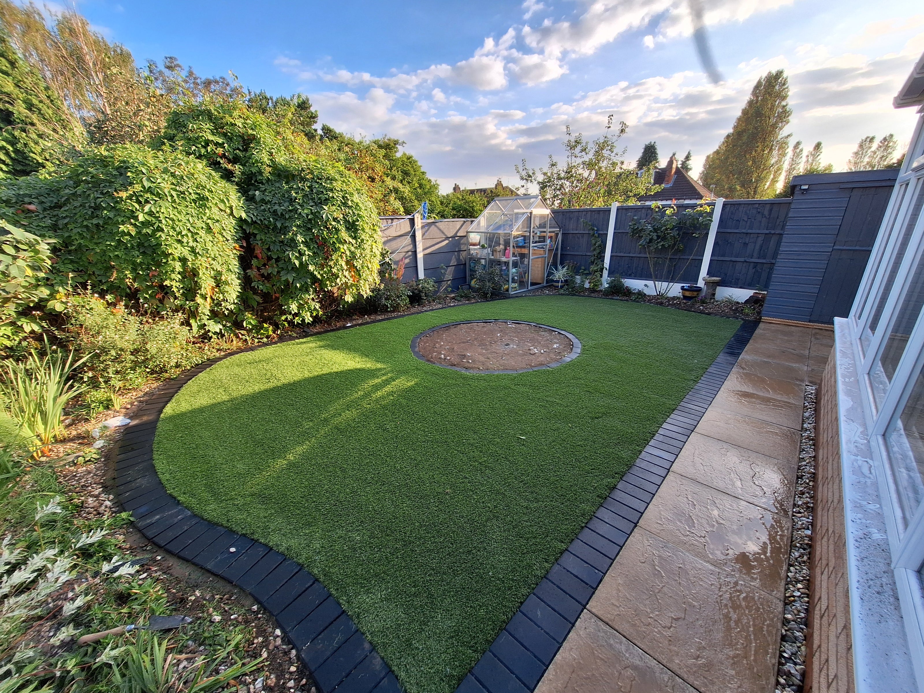 Image shows a fake lawn layed with 50 mm charcoal pavers as a frame with a small circle planting area