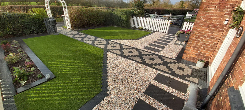 Block paved path in a geometric shape, fake grass either side, with picket fence and hedges in the background