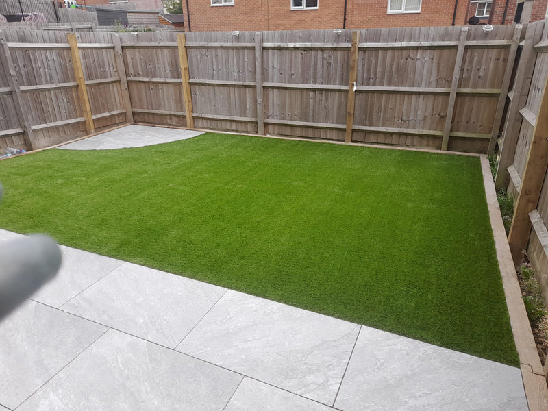 Block paved path in a geometric shape, fake grass either side, with picket fence and hedges in the background
